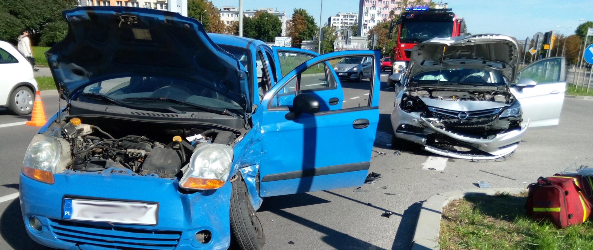 Zdjęcie przedstawia dwa samochody osobowe - jeden niebieski Chevrolet, drugi to srebrny Opel Astra. Jeden z nich - chevrolet ma uszkodzony bok oraz podniesioną do góry maskę - widać skrzywione koło. Drugi samochód stojący za chevroletem ma również podniesioną do góry maskę. Droga jest częściowo zablokowana.