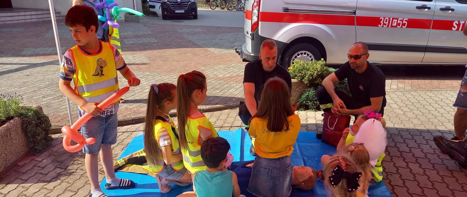 Dwóch strażaków udzielających grupie dzieci instruktażu pierwszej pomocy i resuscytacji krążeniowo oddechowej na fantomie. W tle oznakowany samochód strażacki oraz karetka pogotowia ratunkowego.