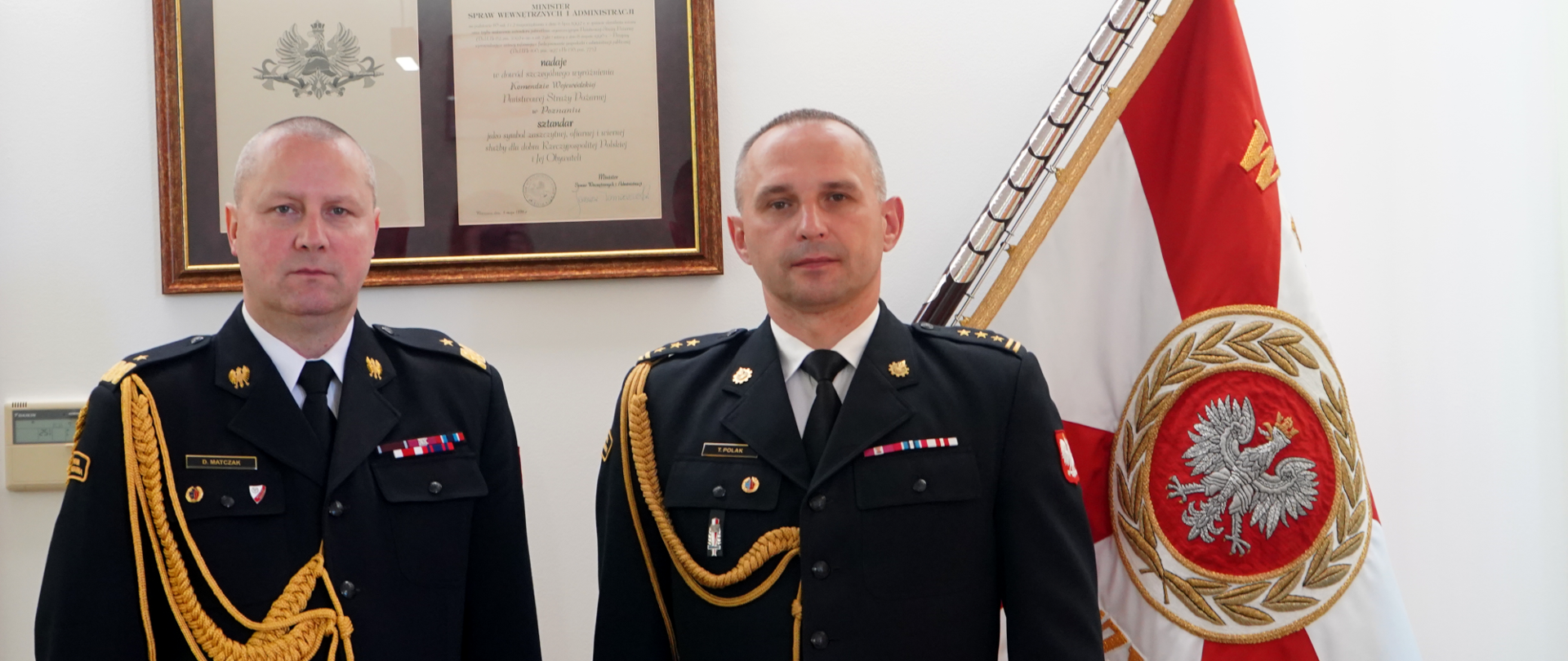 dwóch strażaków w ciemnych mundurach ze złotym sznurem stoi obok siebie, w tle na ścianie obraz oraz sztandar