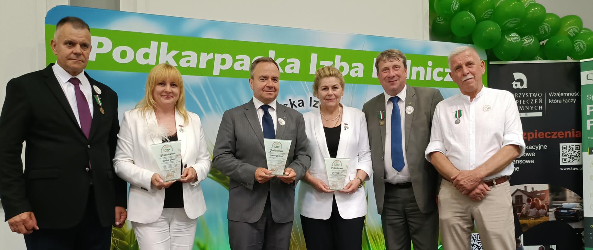 Na scenie podczas uroczystości związanej z Podkarpacką Izbą Rolniczą stoi sześć osób. Trzy osoby w centralnej części zdjęcia trzymają pamiątkowe statuetki otrzymane z okazji jubileuszu. W tle znajduje się duży baner z napisem „Podkarpacka Izba Rolnicza”, zielone balony oraz roll-up partnera wydarzenia. Wszyscy uczestnicy są ubrani w stroje galowe i pozują do wspólnego zdjęcia. Kilka osób ma przypięte odznaczenia na piersi.