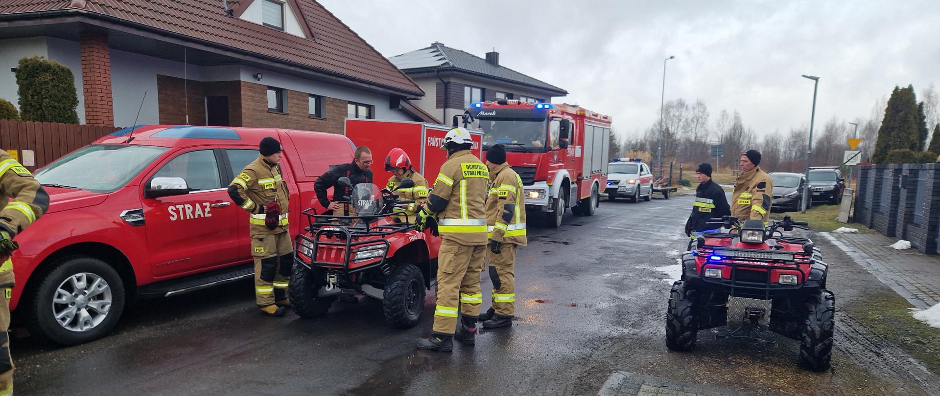 Na zdjęciu druhowie Ochotniczych Straży Pożarnych i funkcjonariusze Państwowej Straży Pożarnej w rejonie koncentracji przy działaniach poszukiwawczych. Strażacy ubrani w ubrania specjalne, obok nich samochody pożarnicze i dwa pojazdy typu quad.
