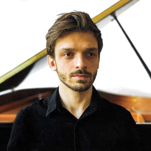 Zdjęcie popiersia młodego mężczyzny z ciemnymi włosami zaczesanymi do góry z brodą. Ubrany w czarną koszulę. Za nim widoczny fragment otwartej klapy czarnego fortepianu.