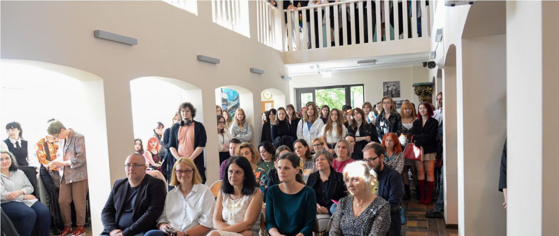 Uroczyste rozpoczęcie roku szkolnego 2022/2023. Nauczyciele siedzący i młodzież w pozycji stojącej zwrócona frontem do robiącego zdjęcie. Pomieszczenie to biały hol szkoły z pasami kolumn po obu stronach podtrzymującymi otwarte półpiętro ograniczone drewnianą balustradą. 