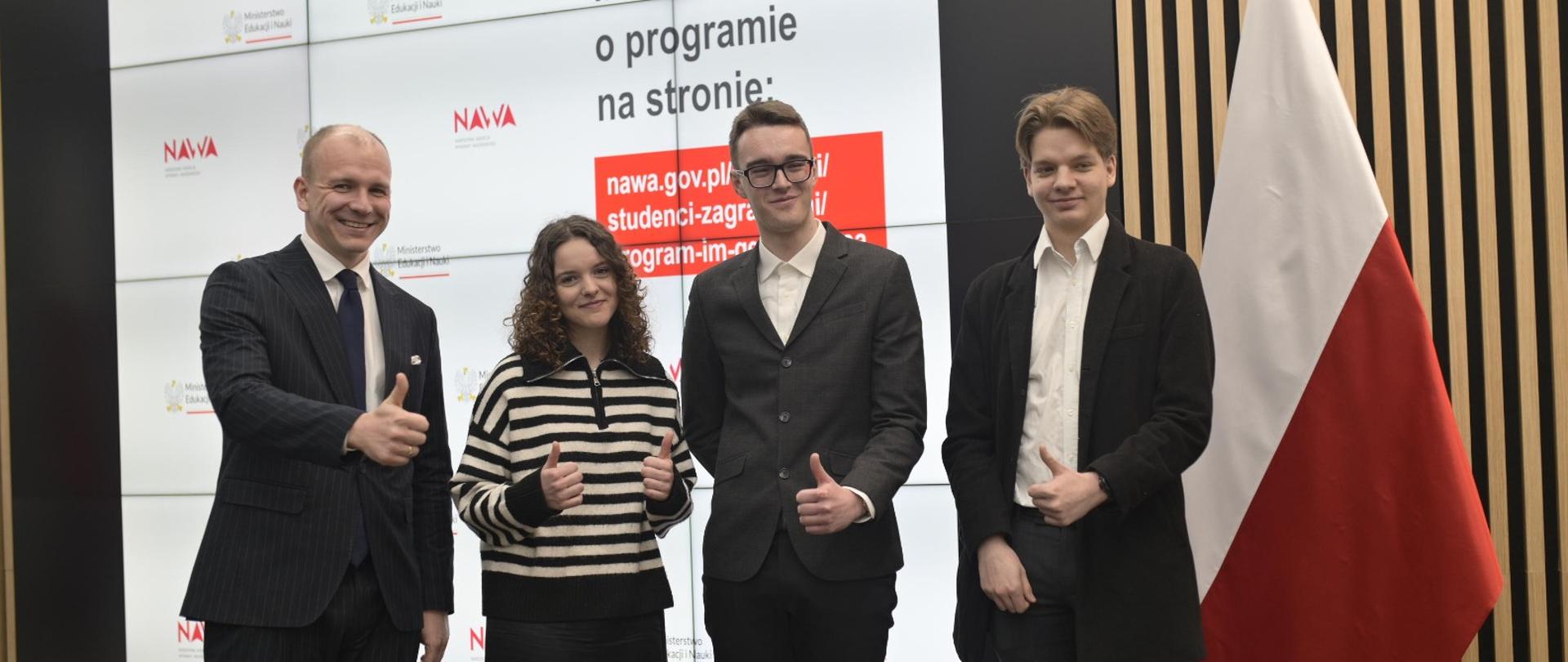 Na tle wielkiego ekranu z napisami NAWA stoi trzech mężczyzn w garniturach i kobieta w swetrze w poziome biało-czarne paski, wszyscy uśmiechają się i pokazują kciuki w górę, za nimi polska flaga.