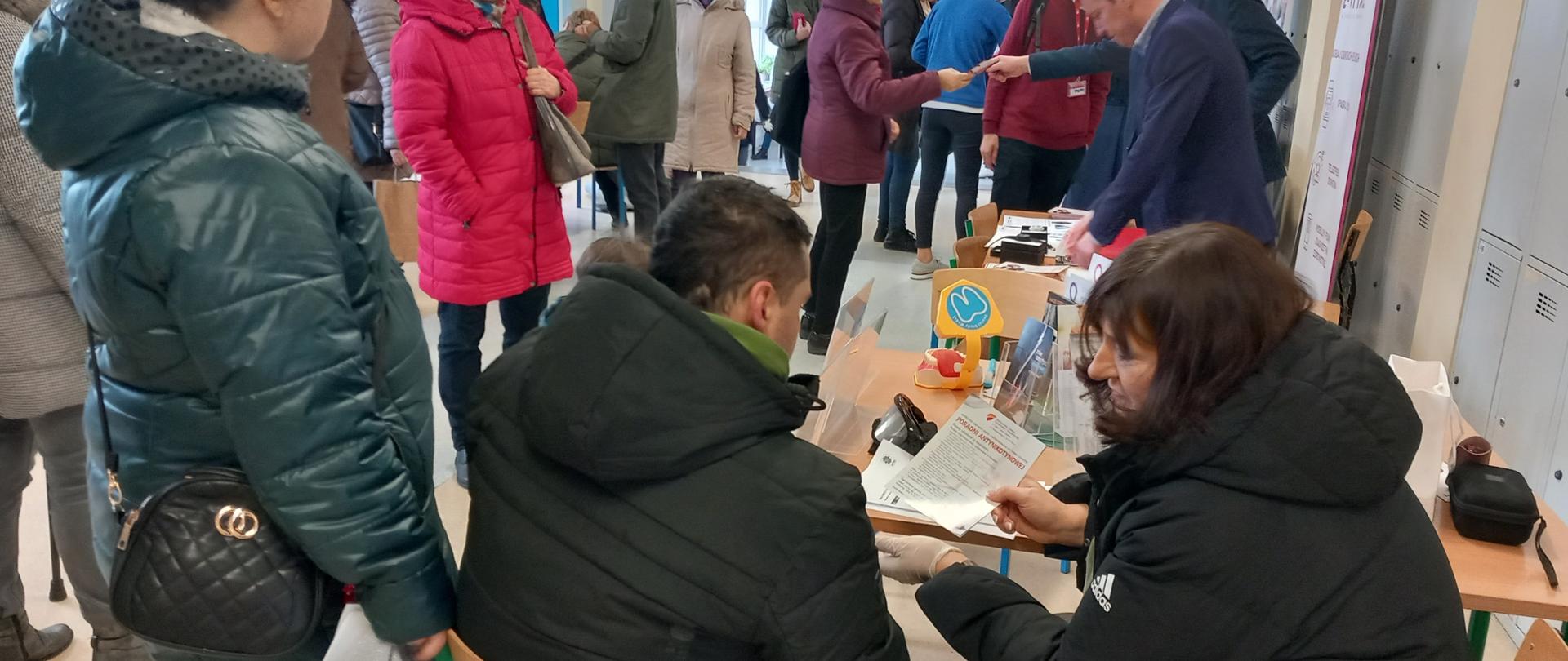 Kobieta siedząca przy stoisku wręcza ulotkę mężczyźnie. W tle wiele osób idących korytarzem. 