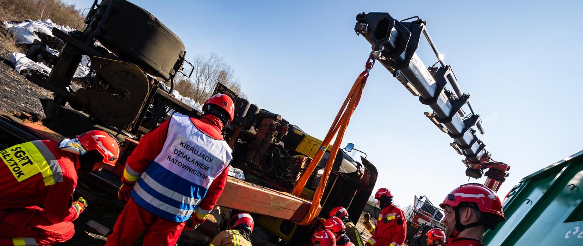 Grupa ratowników technicznych PSP pracuje przy przewróconym wagonie kolejowym, część z nich udziela pomocy osobie na noszach, nadzoruje ich Kierujący Działaniem Ratowniczym.