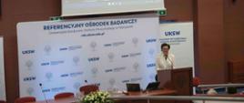 Za drewnianą mównicą stoi kobieta w białej marynarce i mówi do mikrofonu, za nią rozwijany baner z napisem Referencyjny Ośrodek Badawczy, za nim drewniana ściana na której wisi wielki ekran na którym widać ministra Czarnka.
