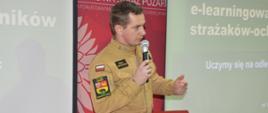 W świetlicy komendy powiatowej PSP, na tle ekranu, na którym wyświetlany jest napis "Platforma e-learningowa dla strażaków-ochotników.Uczymy się na odległość" oraz ścianki reklamowej stoi strażak PSP w ubraniu służbowym koloru piaskowego, który przemawia do siedzących na przeciwko słuchaczy. W prawej ręce trzyma mikrofon.