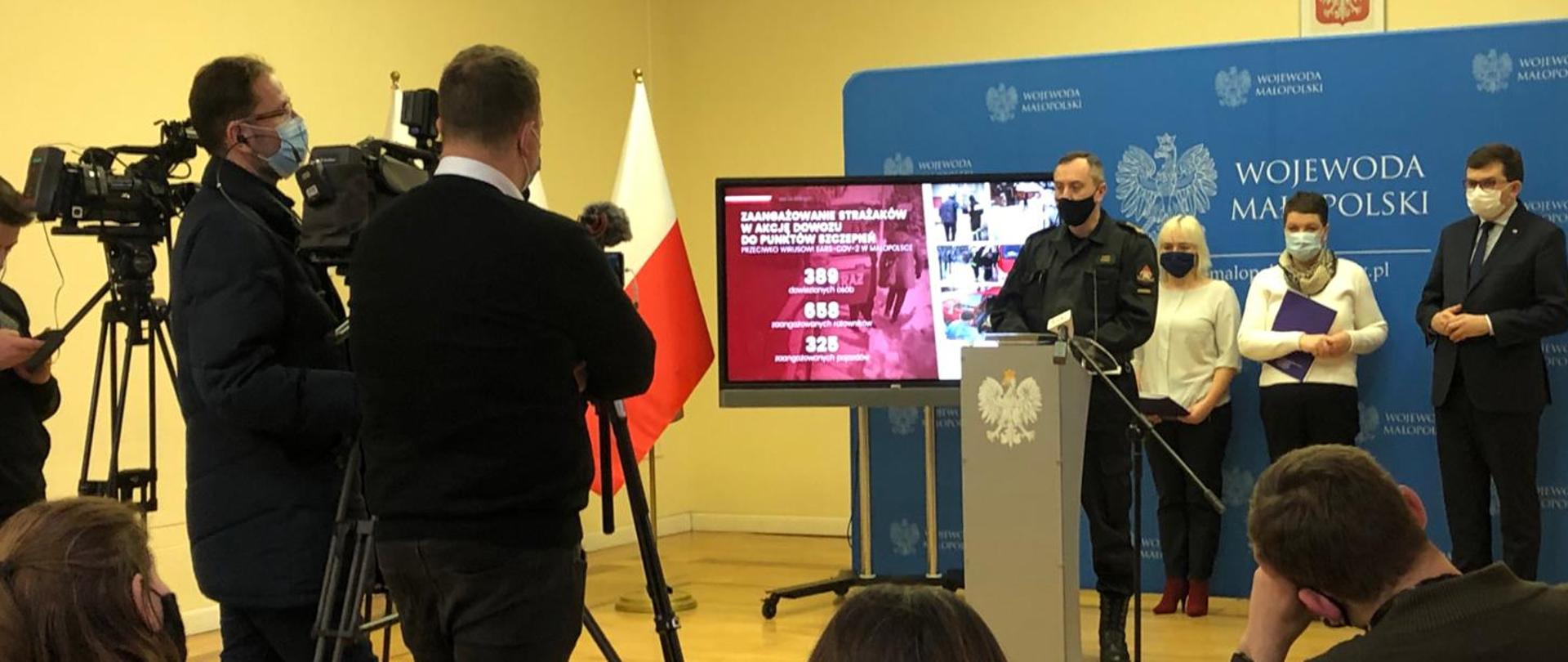 Małopolski Komendant PSP stoi przy mównicy ubrany w mundur dowódczo- sztabowy w czarnej maseczce na twarzy. Za nim stoją dwie kobiety i Wojewoda Małopolski. przed nimi tyłem stoją przy kamerach dziennikarze.