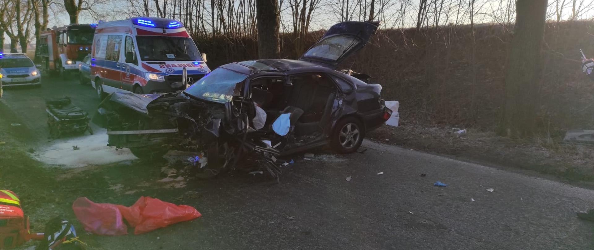 Na zdjęciu widać rozbity pojazd. W tle samochody służb ratowniczych.