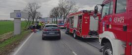 Na jezdni samochody pożarnicze oraz pojazd Policyjny. W oddali widoczne uszkodzone samochody uczestniczące w zdarzeniu. Obok pojazdów działania podejmują strażacy, ratownicy medyczni i policjanci. 