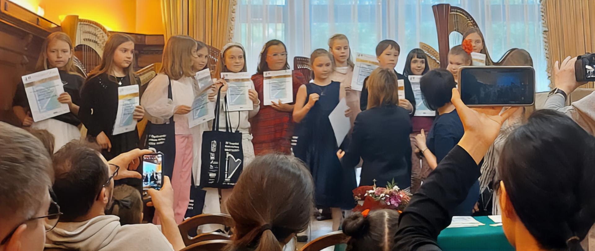 Duża grupa dzieci stoi na scenie, niektóre z nich trzymają w rękach dyplomy, za nimi stoją harfy.