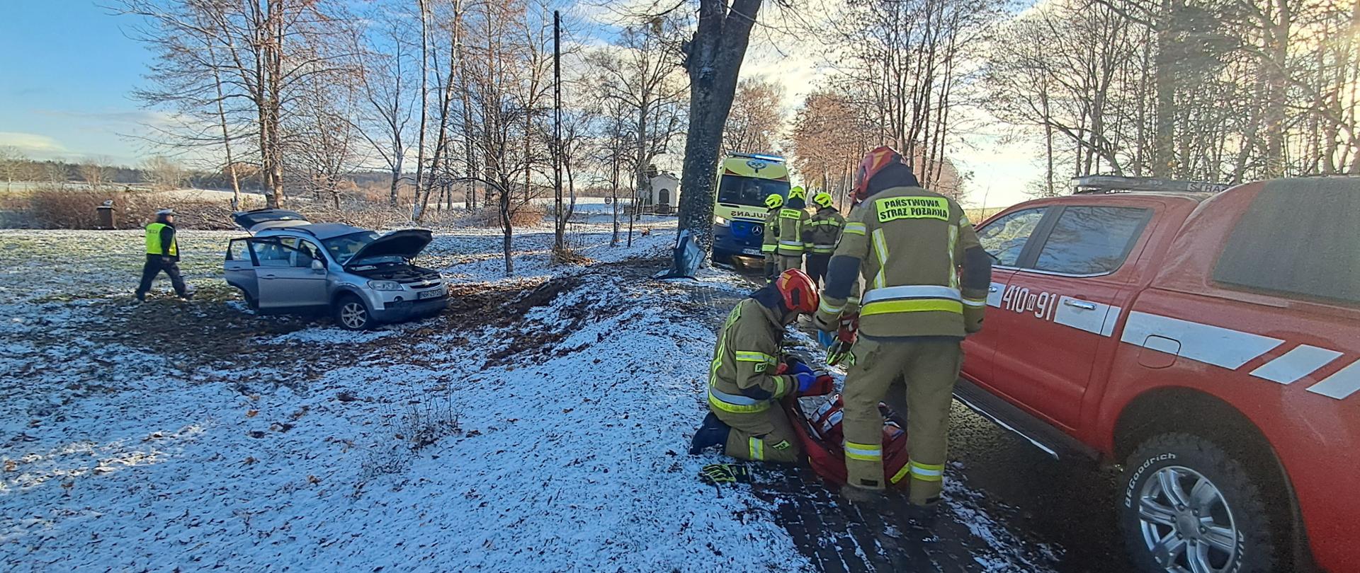 Zdjęcie przedstawia miejsce zdarzenia drogowego, do którego doszło na zaśnieżonej i oblodzonej drodze. Z lewej strony poza drogą samochód osobowy, wypadł z jezdni i uderzył w drzewo. Na miejscu interweniują służby ratownicze – widoczni są strażacy Państwowej Straży Pożarnej udzielający kwalifikowanej pierwszej pomocy osobie poszkodowanej.
W tle znajduje się karetka pogotowia ratunkowego. Warunki atmosferyczne (śnieg, śliska nawierzchnia) mogły przyczynić się do utraty panowania nad pojazdem.
