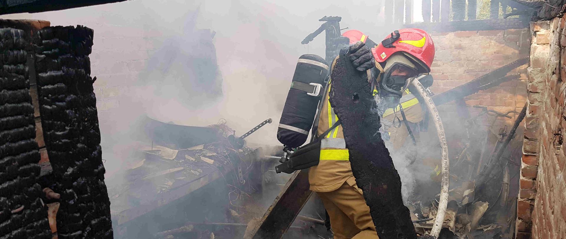 Zdjęcie przedstawia strażaka w ubraniu specjalnym z aparatem ochrony dróg oddechowych, dokonujący rozbiórki budynku. W dłoniach drzyma kawałek spalonej deski. W tle widać spalone wnętrze budynku. 