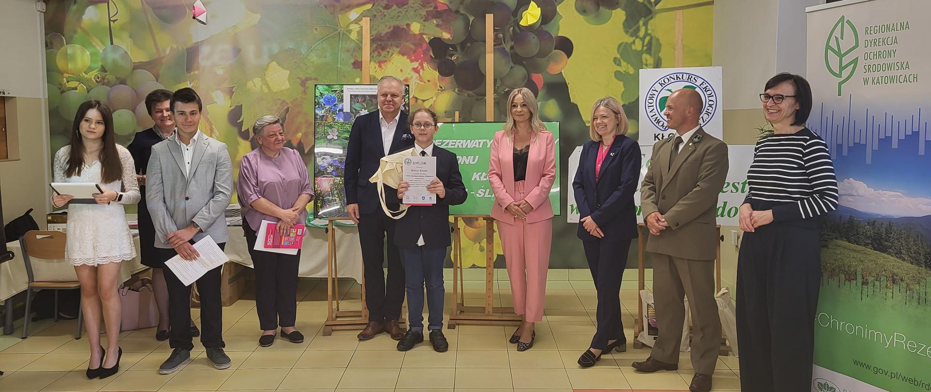 Stojący w półkolu ludzie z uczniem (laureatem) po środku. Uczeń trzyma nagrodę