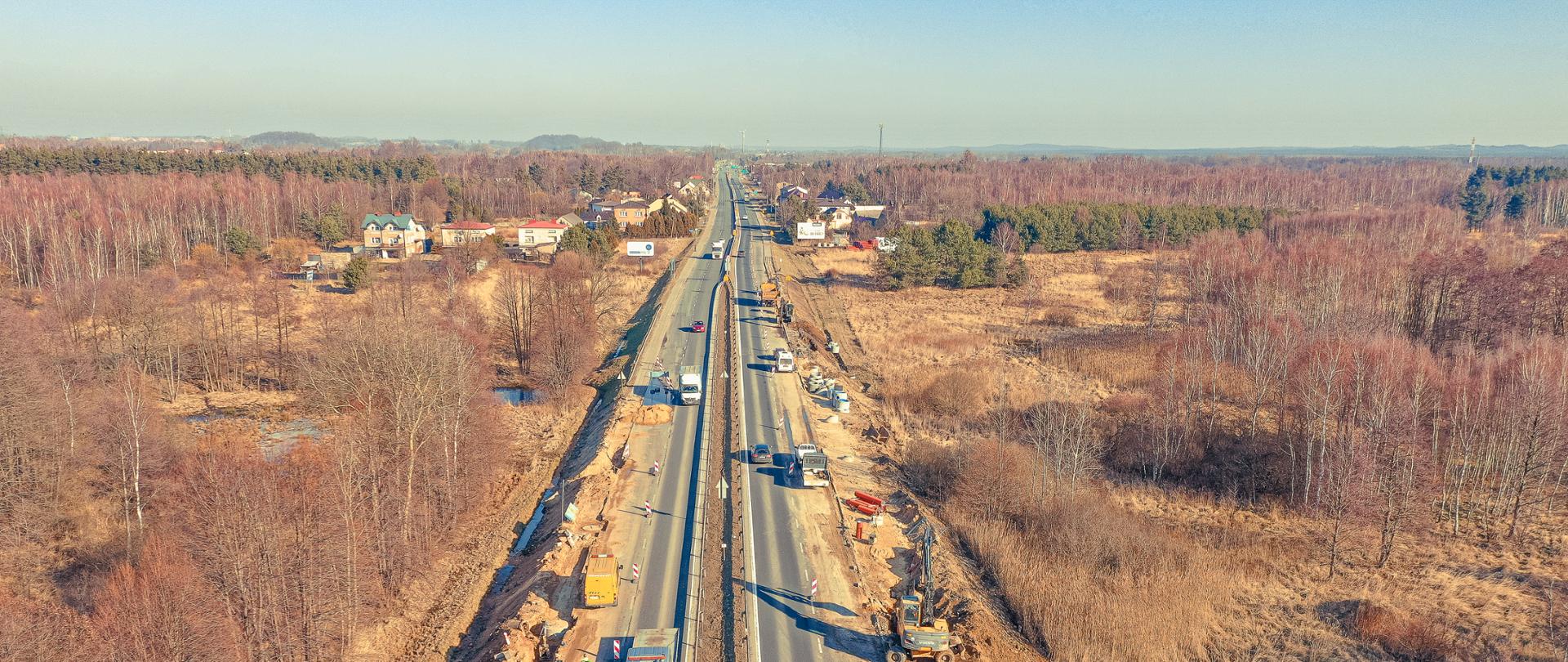 Fotografia lotnicza dwujezdniowej drogi Krajowej 91. Fotografia przedstawia centralnie położona dwujezdniową drogę krajową. Otoczenie drogi słabo zurbanizowane, liczne tereny leśne. Na obu jezdniach wyłączono z ruchu prawe pasy, na których odbywają się prace przy budowie kanalizacji odwodnienia drogi.
