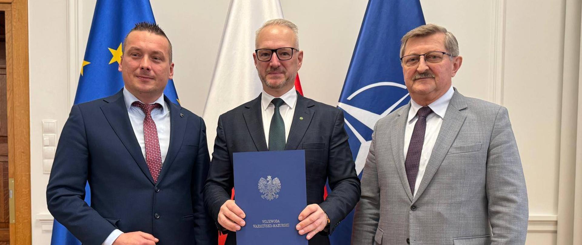 Trzy osoby stoją i trzymają w ręku teczkę za nimi znajdują się flagi. Flaga Unii Europejskiej, Flaga Polski i Flaga Nato