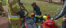 Zdjęcie przedstawia strażaków udzielających kwalifikowanej pierwszej pomocy osobie poszkodowanej leżącej na desce ortopedycznej, wyniesionej z budynku objętego pożarem. 