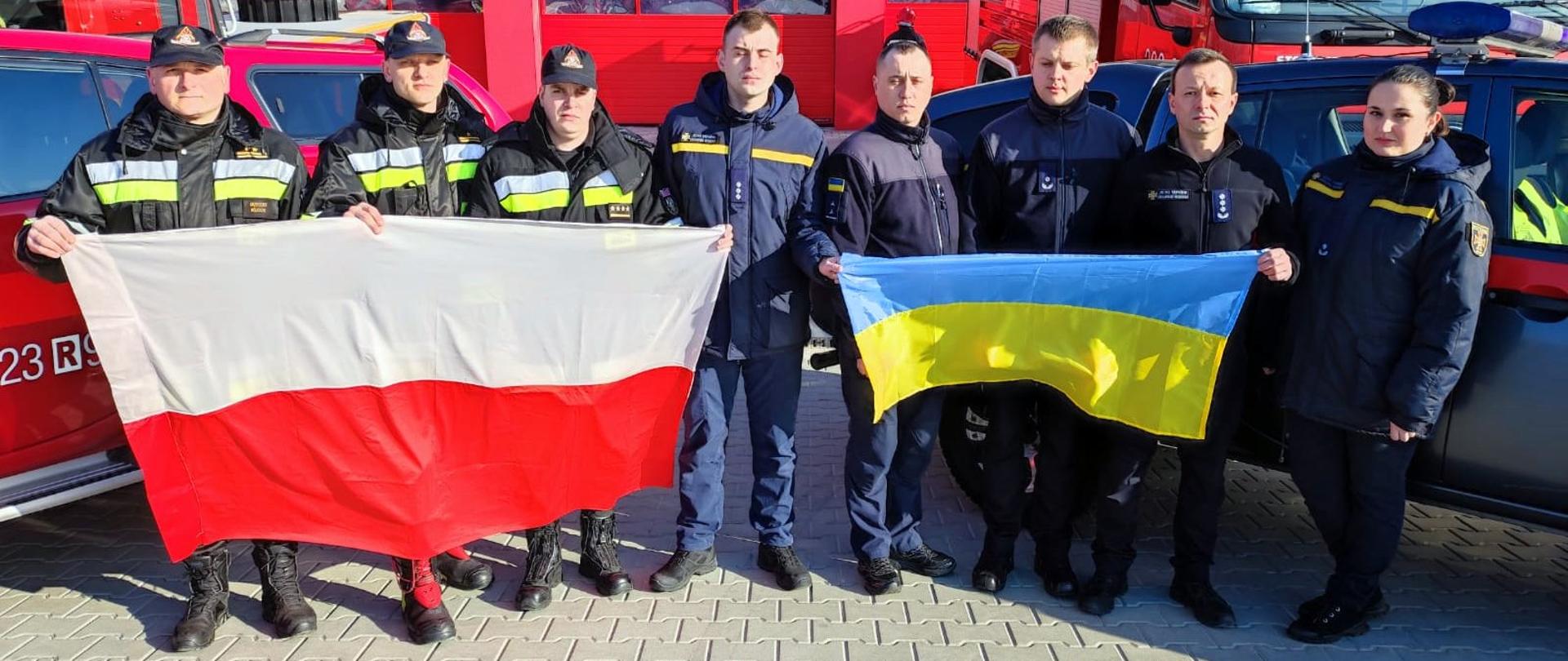 Zdjęcie zrobione przed remizą strażacką. Strażacy ustawieni na uroczystej zbiórce, ubrani w umundurowanie specjalne. W tle ustawione samochody strażackie.