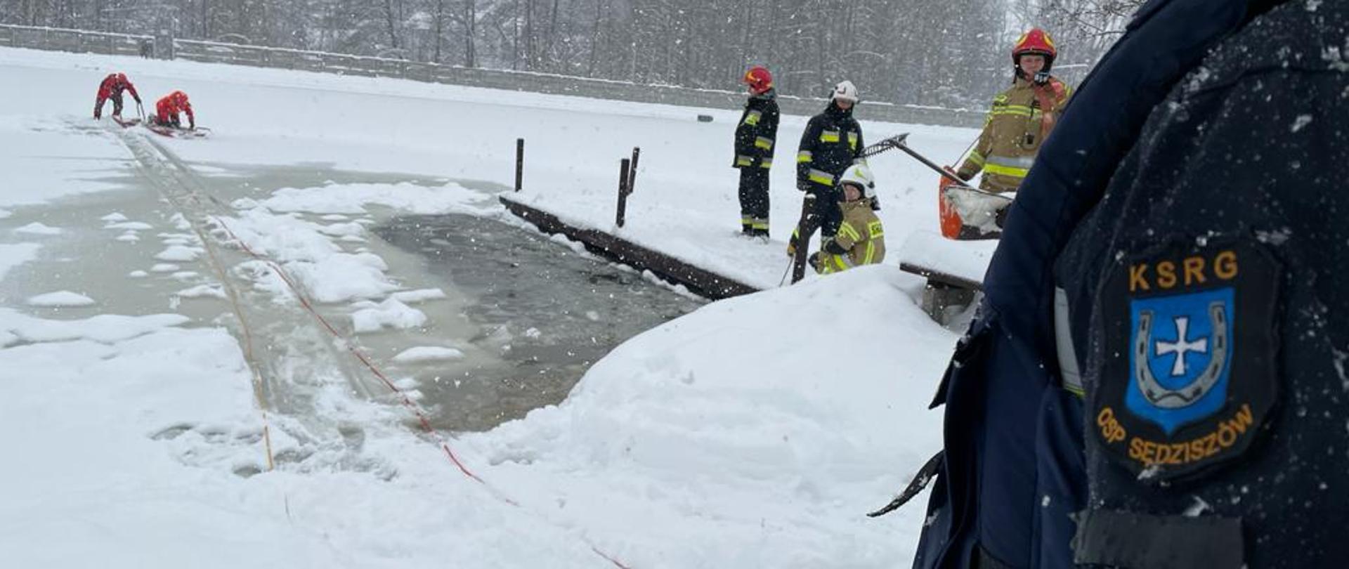 Dwóch strażaków w ubraniach wypornościowych oraz kamizelkach asekuracyjnych w tle na saniach lodowych. Mężczyźni mają założone na głowach kaski ochronne. Znajdują się oni na środku zalewu miejskiego w Sędziszowie, który pokryty jest lodem. Po prawej kilku strażaków w umundurowaniu specjalnym. Na pierwszym planie lewe ramię strażaka ratownika OSP z napisem KSRG OSP Sędziszów.