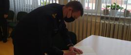 W sali konferencyjnej komendy komendant powiatowy składa podpis na dokumencie. Strażak ubrany w czarny mundur wyjściowy.