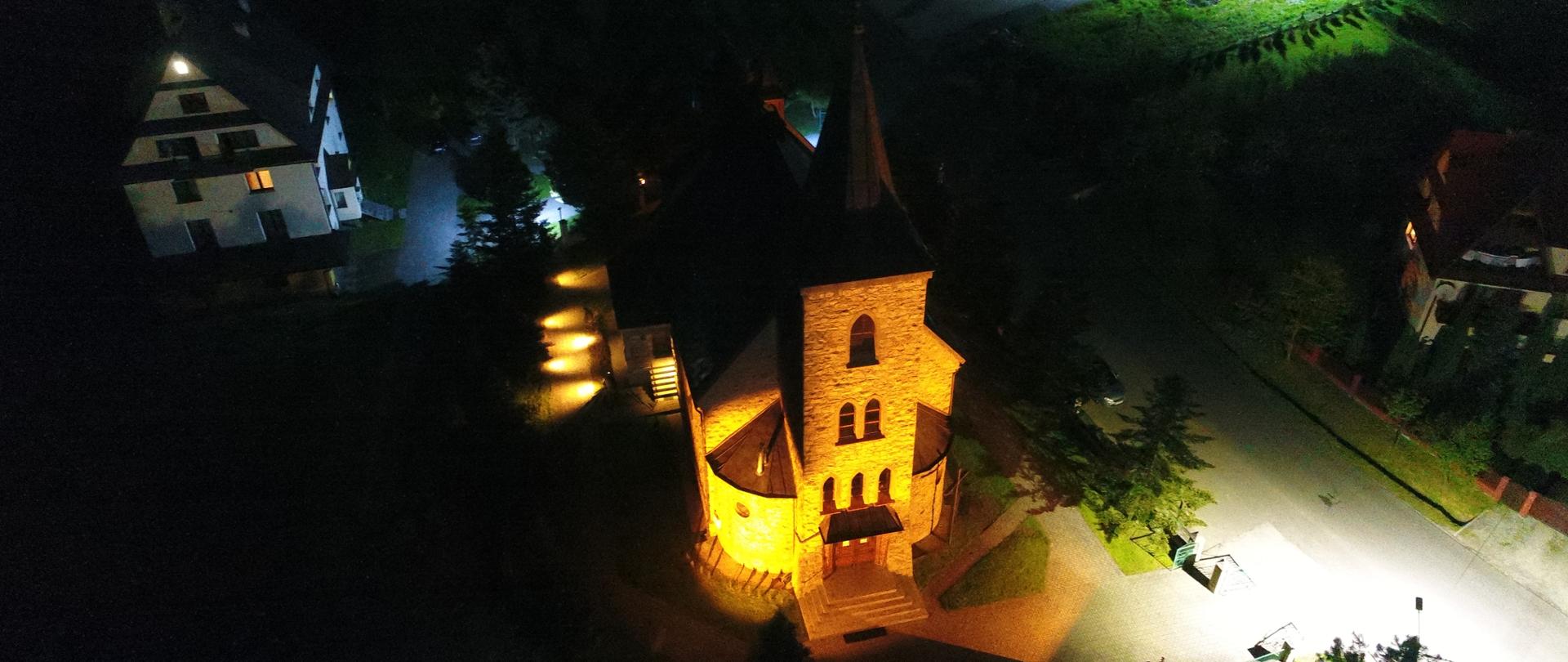 Widok z lotu ptaka na oświetlony pomarańczowym światłem kościół. Część budynku nie jest oświetlona. Z prawej strony widać fragment drogi z intensywnym, białym oświetleniem.
