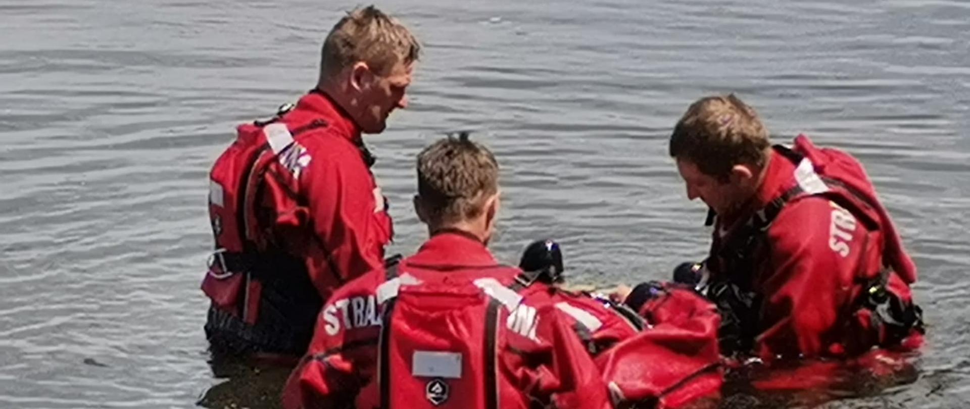 Na zdjęciu widoczny akwen. W wodzie znajduje się czterech strażaków w czerwonych suchych skafandrach nurkowych z napisem straż. Trzech z nich stoi i podtrzymuje czwartego, unoszącego się na wodzie.
