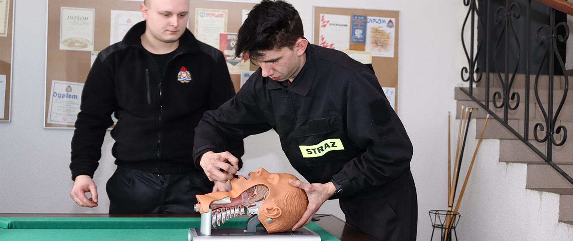 na stole leży fantom do nauki intubacji ratownik osp ubrany w ubranie koszarowe zakłada manekinowi rurkę gardłowo-ustną drugi ratownik ubrany w ubranie koszarowe przygląda się koledze

