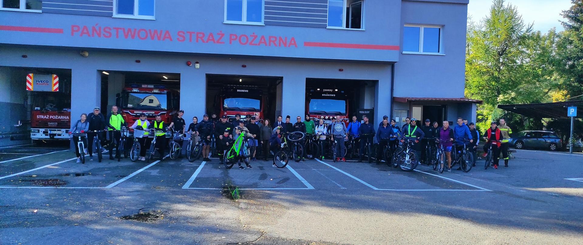 Przed budynkiem straż pożarnej na tle garaży z samochodami pożarniczymi stoi grupa kilkudziesięciu osób z rowerami. 
