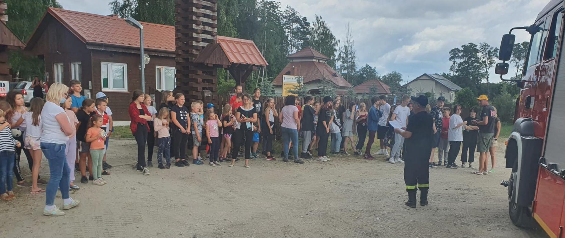 Na zdjęciu widać grupę dzieci i opiekunów stojących przed ośrodkiem wczasowym a przed nimi wóz strażacko oraz przemawiający do nich strażak.