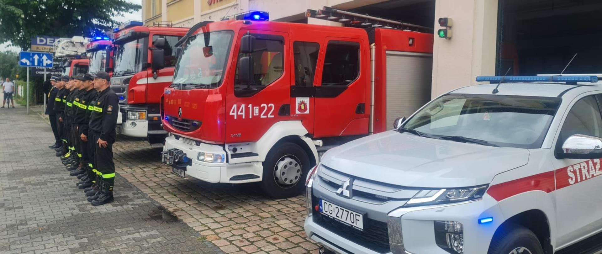 Zbiórka strażaków w ciemnych mundurach przed czerwonymi pojazdami strażackimi, które częściowo wystają przed budynek. Strażak z lewej oddaje honor do rogatywki.