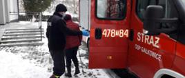 Zdjęcie przedstawia samochód strażacki na tle wejścia do budynku Niepublicznego Zakładu Opieki Zdrowotnej w Dwikozach. Na zdjęciu widać jak strażak pomaga wyjść osobie starszej z samochodu strażackiego. 
