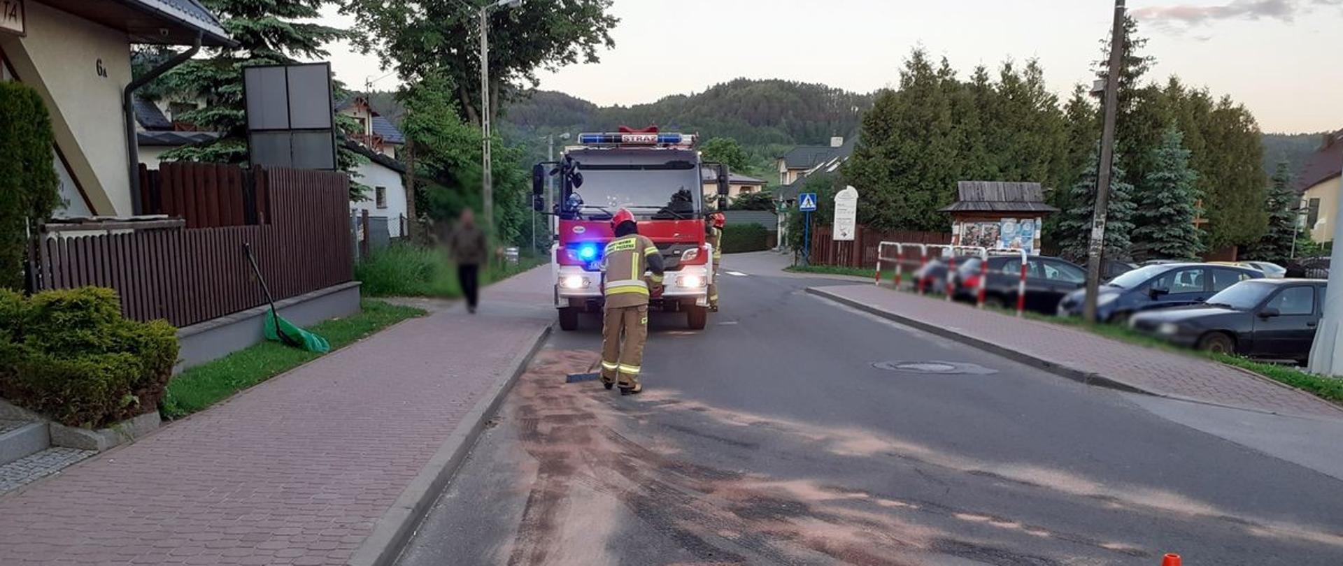 Strażak przy pomocy miotły rozprowadza sorbent na drodze.W tle stoi samochód pożarniczy.Obok drogi na parkingu stoją samochody.