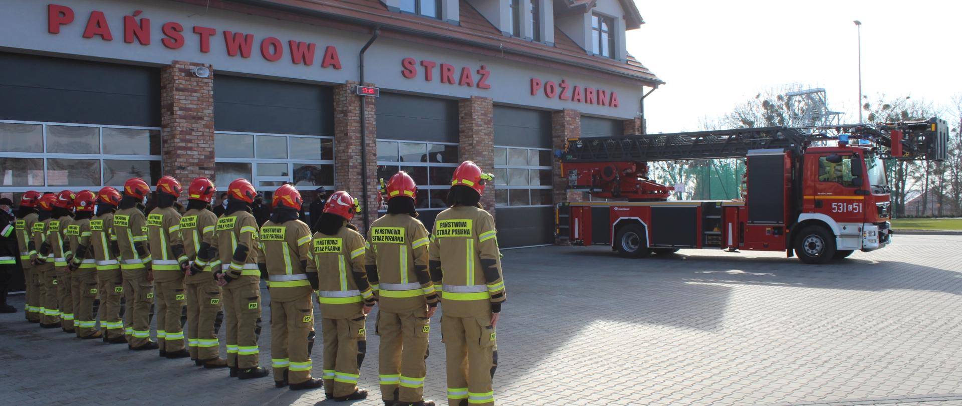 Na placu przed budynkiem Komendy Powiatowej Państwowej Straży Pożarnej w Świeciu po lewej stronie zdjęcia 13 strażaków ubranych w ubrania specjalne oraz hełmy stoi tyłem w szeregu. Po prawej stronie zdjęcia w tle widać nowy samochód pożarniczy z drabiną mechaniczną.
