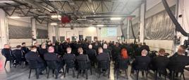 Na zdjęciu widać strażaków w mundurach wyjściowych siedzących na krzesłach. Skierowani są w kierunku rzutnika na którym są wyświetlane informacje dot. odprawy. Zebranie zorganizowane jest wewnątrz garażu Komendy Powiatowej.