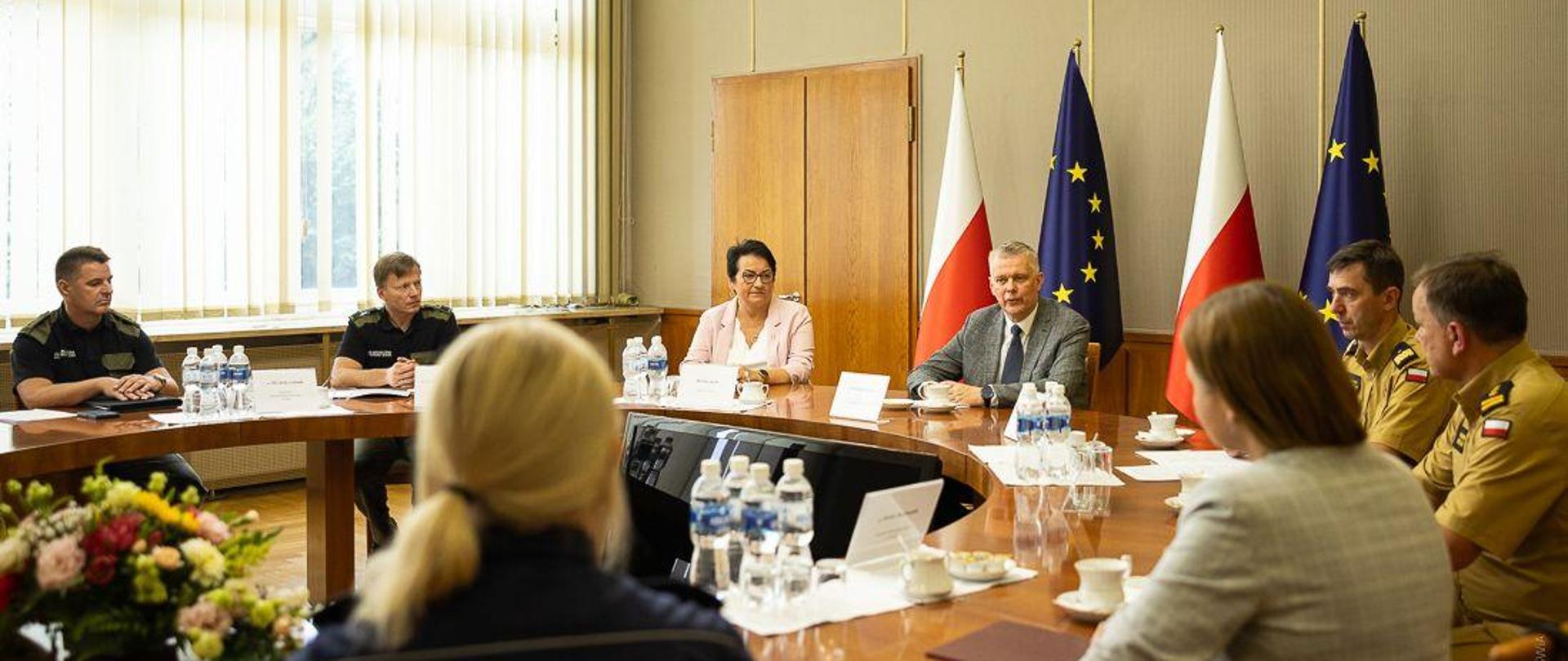 Grupa osób na czele z ministrem Siemoniakiem siedzi przy okrągłym stole. W tle flagi polskie i unijne 
