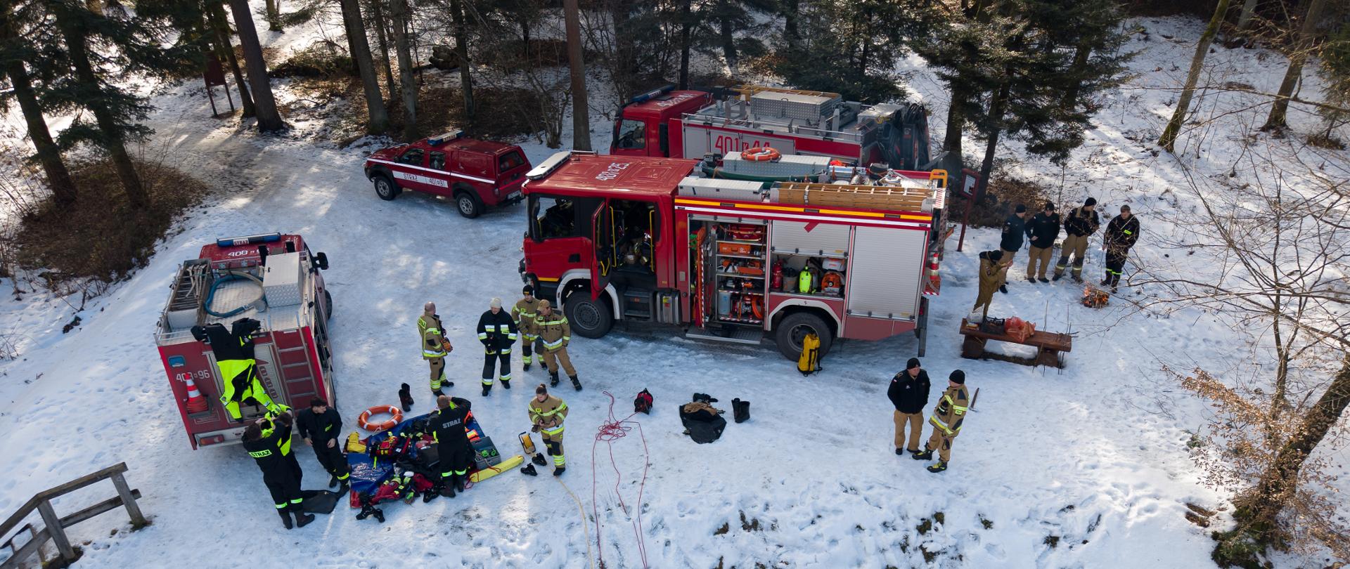 Zdjęcie przedstawia ćwiczenia z zakresu ratownictwa lodowego. Na zdjęciu widać stojące na brzegu stawu cztery samochody pożarnicze w kolorze czerwonym. . Obok pojazdów stoją strażacy ubrani w ubrania specjalne w kolorze piaskowym. Na ziemi obok pojazdów na macie sprzętowej leży sprzęt do ratownictwa wodno-lodowego. Na zamarzniętej wodzie na zbiorniku stoją sanie lodowe a przy nim strażak asekurowany na lince ratowniczej.