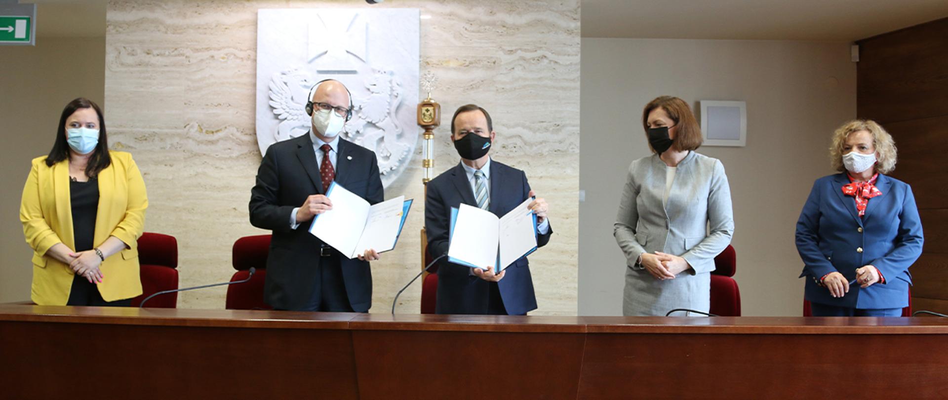 W pomieszczeniu pięć osób stoi za długim stołem. Pierwsza od lewej wiceminister Małgorzata Jarosińska-Jedynak. Dwóch mężczyzn prezentuje dokumenty.