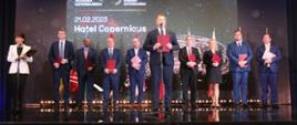 Kilka trzymających czerwone teczki osób stoi na błyszczącej podłodze, za nimi na ścianie napis 21.02.2023 Hotel Copernicus, przed nimi stoi minister Czarnek i mówi do mikrofonu na stojaku.