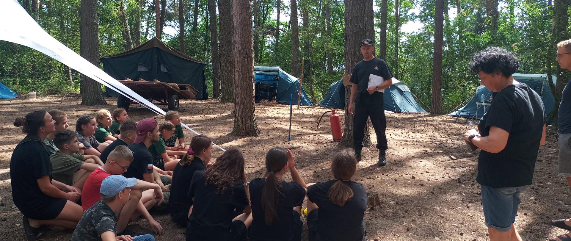 Młodzież siedzi na zmiemi w lesie obok rozstawionych bamiotów. Dorośli stoją.