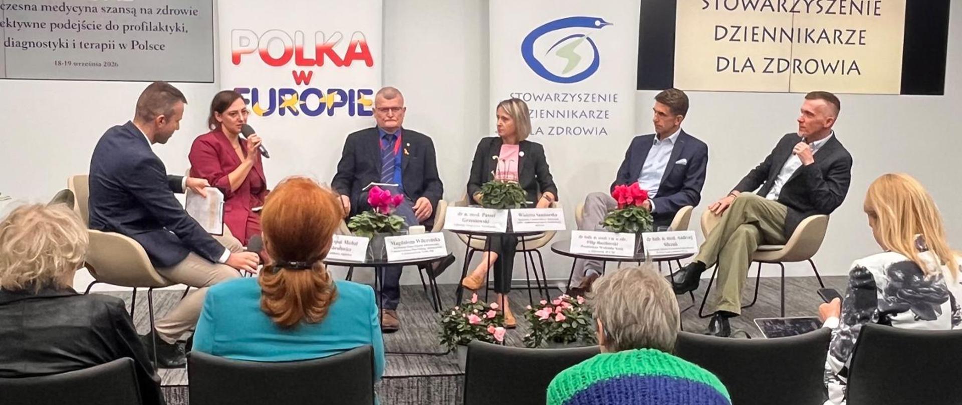 Panel dyskusyjny podczas wydarzenia poświęconego zdrowiu. Na scenie siedzi pięcioro panelistów oraz moderator, a przed nimi znajdują się tabliczki z nazwiskami. W tle widoczne są logotypy i materiały organizatorów, w tym Stowarzyszenia Dziennikarze dla Zdrowia.