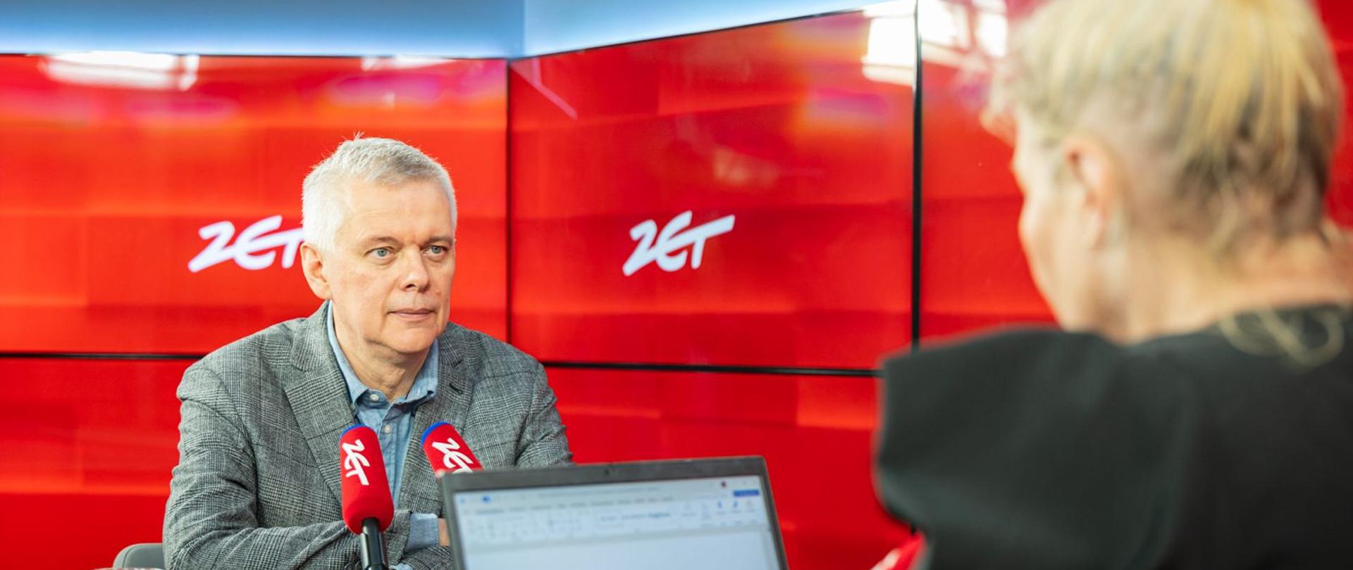 Minister Tomasz Siemoniak siedzi przy stole podczas wywiadu. Przed nim znajdują się dwa czerwone mikrofony z białym logo oraz szklanka wody. Ubrany jest w szarą marynarkę w kratę i niebieską koszulę. W tle widoczne są czerwone ekrany z białymi znakami „zet”. Na pierwszym planie, częściowo zasłaniając kadr, znajduje się osoba siedząca tyłem z laptopem. Scena odbywa się w studiu.