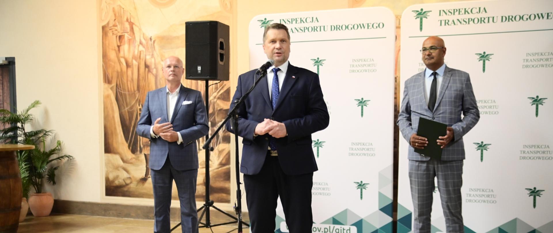 Trzech mężczyzn w garniturach stoi na tle dużego baneru z napisem Inspekcja Transportu Drogowego. Jeden z nich stoi przed mikrofonem i przemawia.