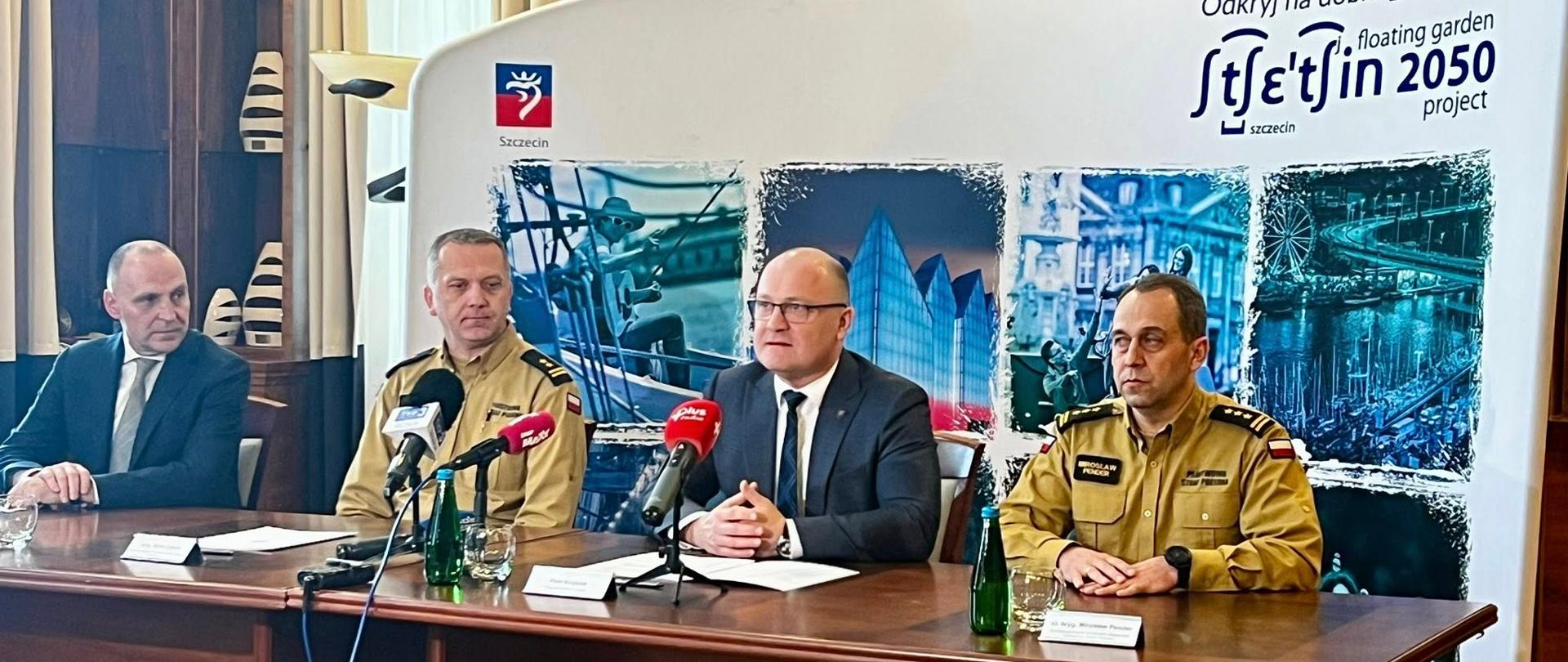 Na zdjęciu widać czterech mężczyzn siedzących za długim drewnianym stołem. Za nimi znajduje się białe tło z napisem "Odkryj na dobre... Szczecin floating garden in 2050 project" oraz logo miasta Szczecin. Na tle widnieją również trzy zdjęcia przedstawiające projekty związane z "floating garden".
Dwóch mężczyzn siedzących po lewej stronie stołu ubranych jest w ciemne garnitury. Mężczyzna siedzący najbardziej po lewej ma na sobie czarny garnitur, białą koszulę i ciemny krawat. Mężczyzna siedzący obok niego ma na sobie beżowy mundur z ciemnymi naramiennikami i guzikami.
Dwóch mężczyzn siedzących po prawej stronie stołu ubranych jest w podobne mundury w kolorze piaskowym z ciemnymi naramiennikami i naszywkami. Mężczyzna siedzący najbliżej środka ma na sobie ciemny garnitur, białą koszulę i czerwony krawat.
Na stole przed każdym z mężczyzn stoi przezroczysta butelka wody i leży biała kartka. Przed środkowymi mężczyznami stoją również mikrofony. Stół jest ustawiony na tle drewnianej ściany z półkami. Po lewej stronie widoczna jest jasna lampa stojąca o nietypowym kształcie.
