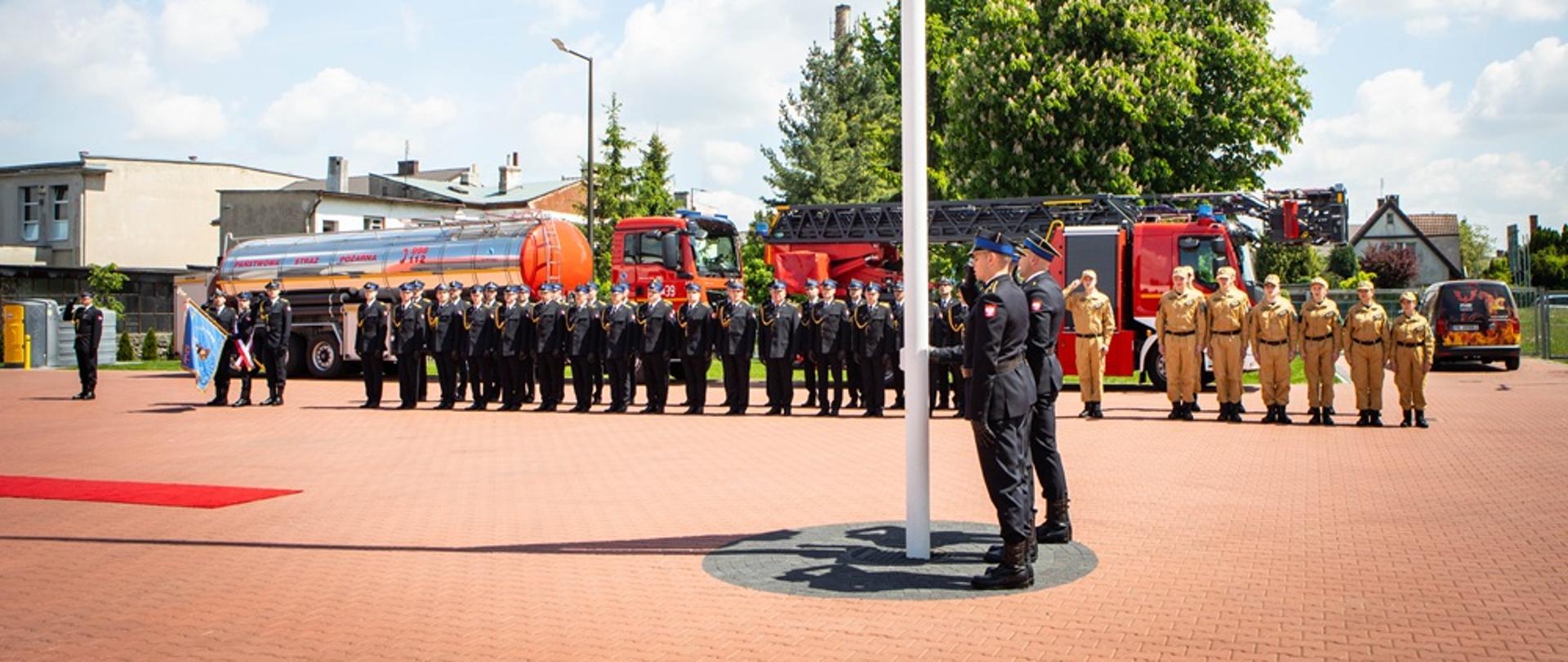 Strażacy w ubiorze wyjściowym podnoszą flagę państwową. 