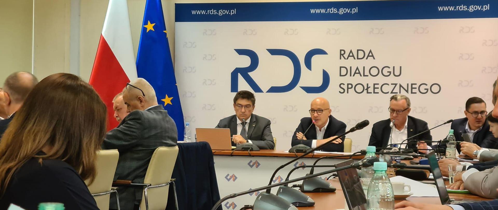 Uczestnicy spotkania siedzą wokół okrągłego stołu. W tle ścianka z napisem Rada Dialogu Społecznego i flagi. 