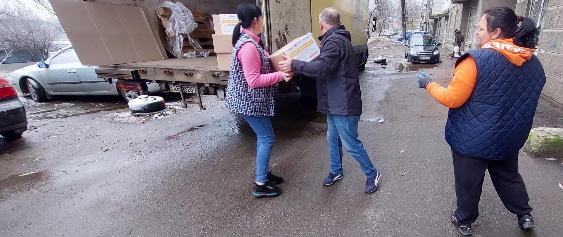 Kobieta i mężczyzna wyciągają paczki z ciężarówki zaparkowanej na ulicy i podają je kolejnej kobiecie.