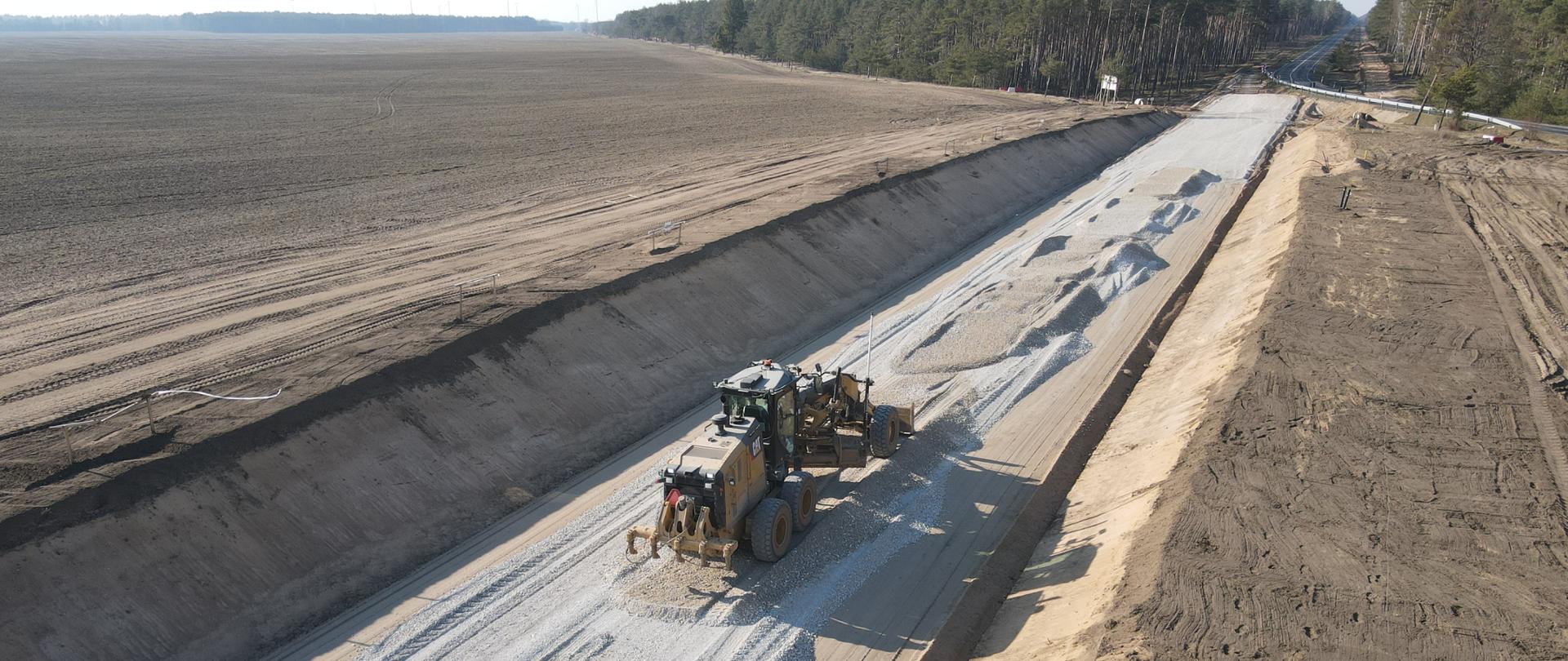 Zdjęcie przedstawia budowę obwodnicy Brzezia w ciągu drogi krajowej nr 25. Na pierwszym planie równiarka przygotowuje konstrukcję drogi. W oddali widać obecny przebieg DK25.