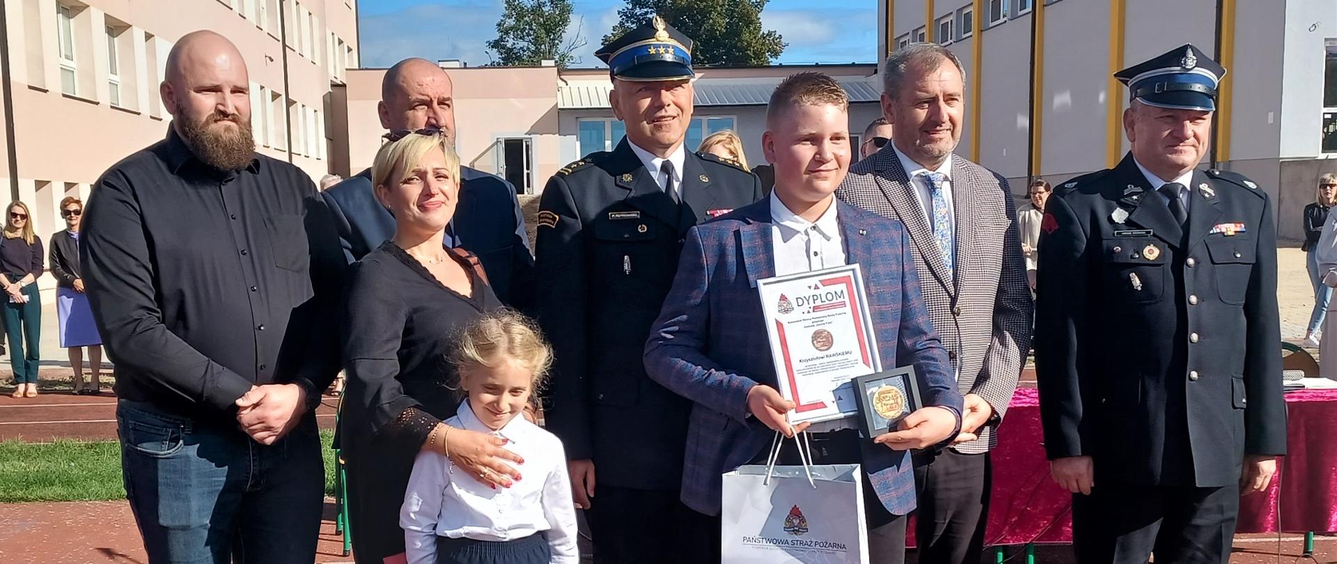 Grupa ludzi, a w ich gronie mały bohater, który trzyma w reku odznakę i dyplom. Za nimi strażacy w mundurach wyjściowych, rodzice, rodzeństwo. W tle budynek szkoły.