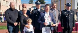 Grupa ludzi, a w ich gronie mały bohater, który trzyma w reku odznakę i dyplom. Za nimi strażacy w mundurach wyjściowych, rodzice, rodzeństwo. W tle budynek szkoły.